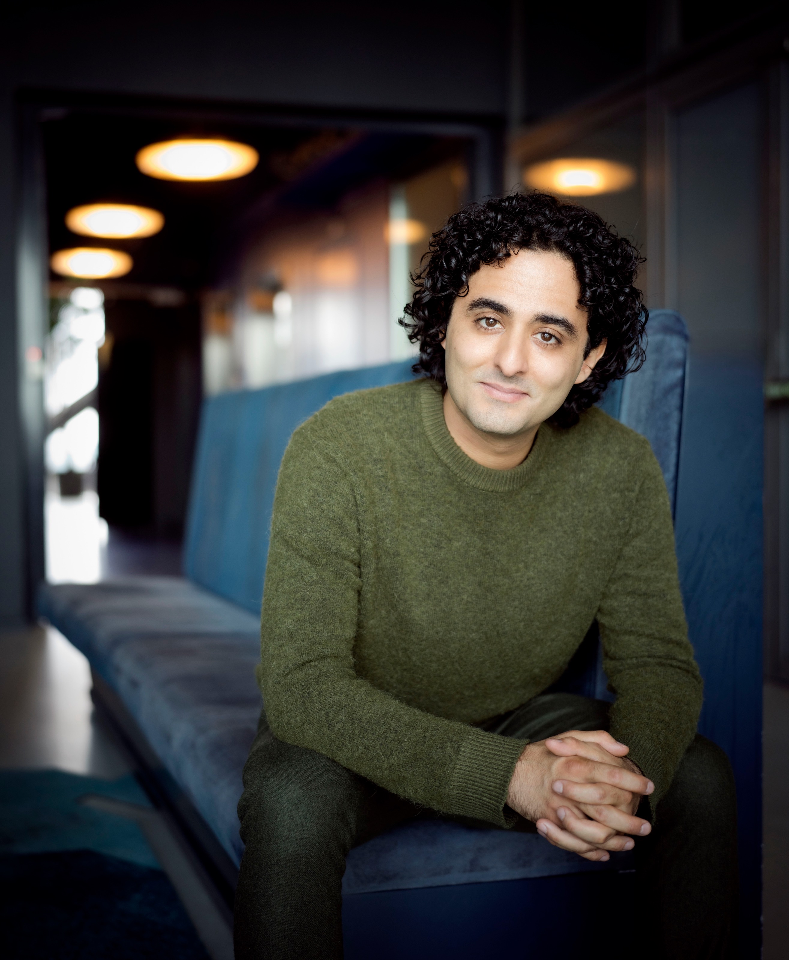 Kerem Hasan seated on a blue bench in an olive green sweater